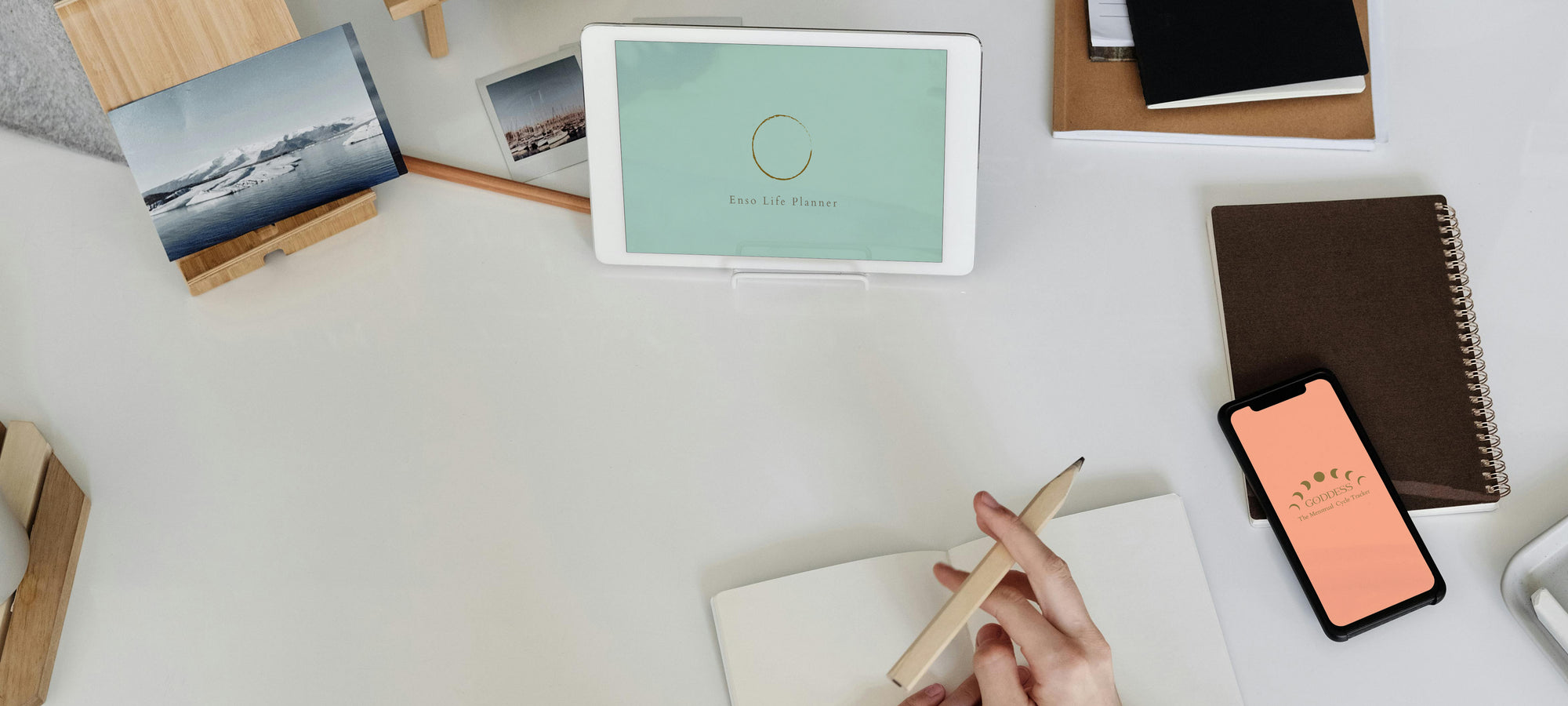 Enso Life Planner on ipad, Goddess tracker on phone screen on aesthetically laid out desk, a hand holds a pencil above a notebook